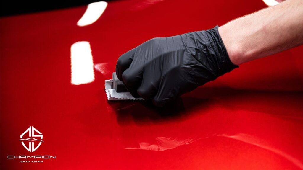 A gloved hand applies ceramic coating with a microfiber applicator to a glossy red car hood, reflecting bright white lights. The Champion Auto Salon logo is in the lower left corner.
