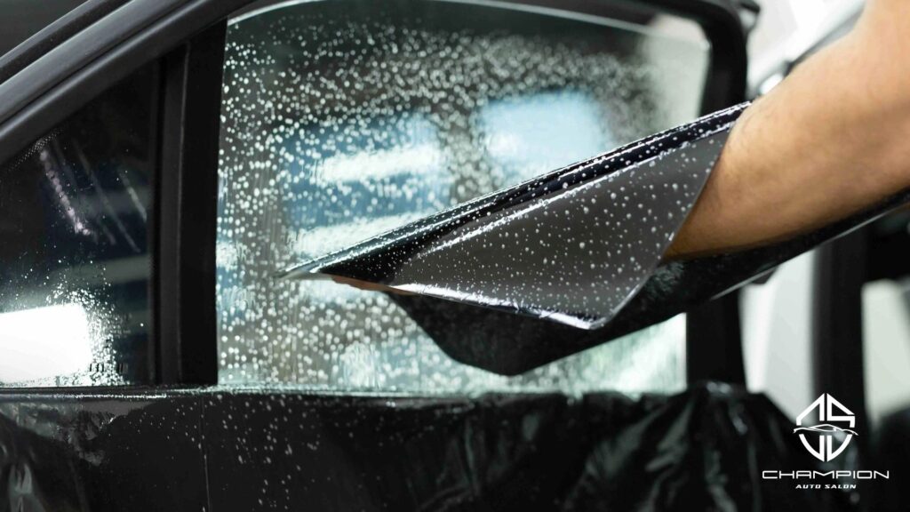 A tint film sheet is carefully peeled and positioned on a soapy car window, ready for installation. Champion Auto Salon logo is visible in the corner.