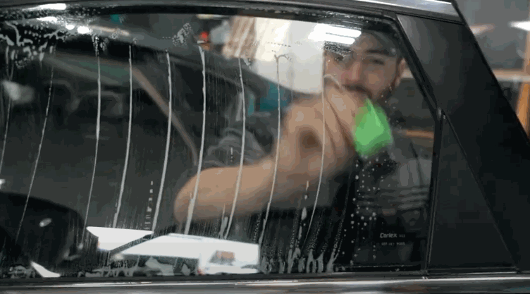 A man scrubs the inside of a car window using a green squeegee-like tool, removing cleaning solution.