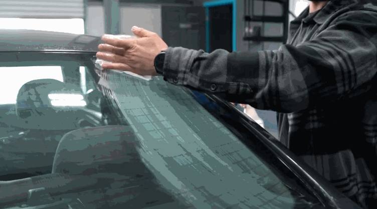 A person smooths a sheet of tint film onto a car’s windshield, aligning it carefully with their hands inside a workshop.