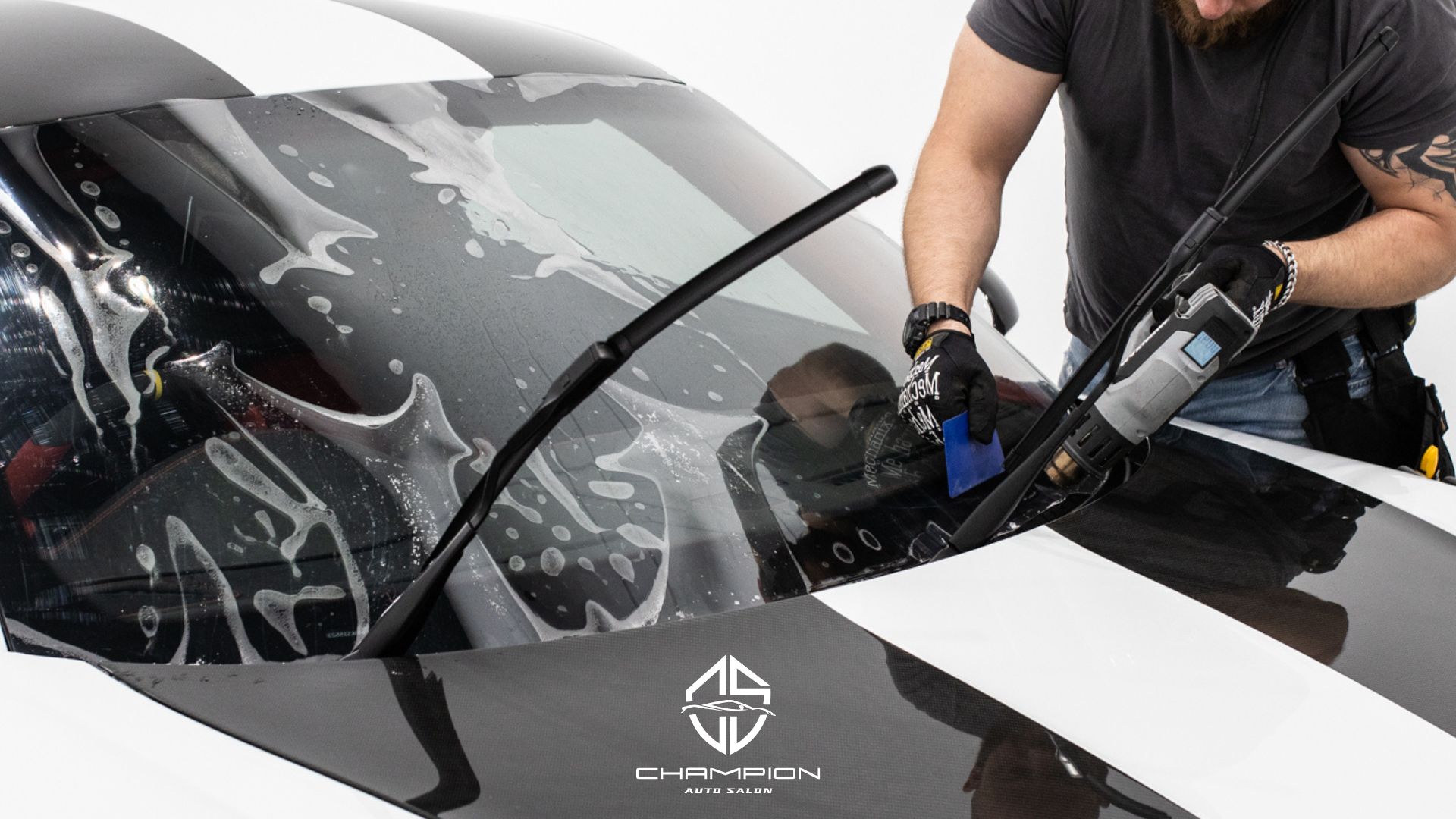 A technician uses a heat gun and squeegee to apply window tint to a sports car windshield covered in soapy water.