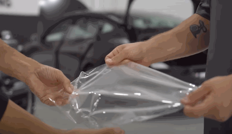 Two people stretch a piece of clear paint protection film to demonstrate its flexibility and durability before installation.