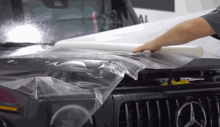 A technician begins laying a clear paint protection film sheet over the hood of a black Mercedes-Benz G-Class SUV.
