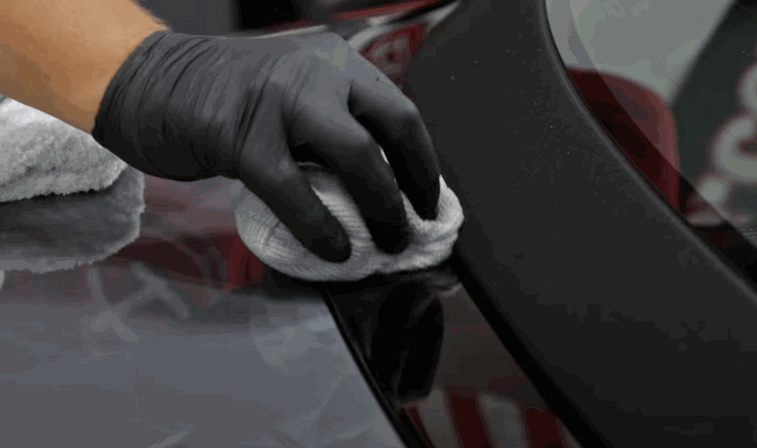 A gloved hand uses a microfiber towel to buff off excess ceramic coating from a black vehicle surface.