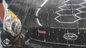 Water beads on the hood of a black Subaru WRX STI, showing the hydrophobic effect of ceramic coating. Champion Auto Salon logo is in the corner.