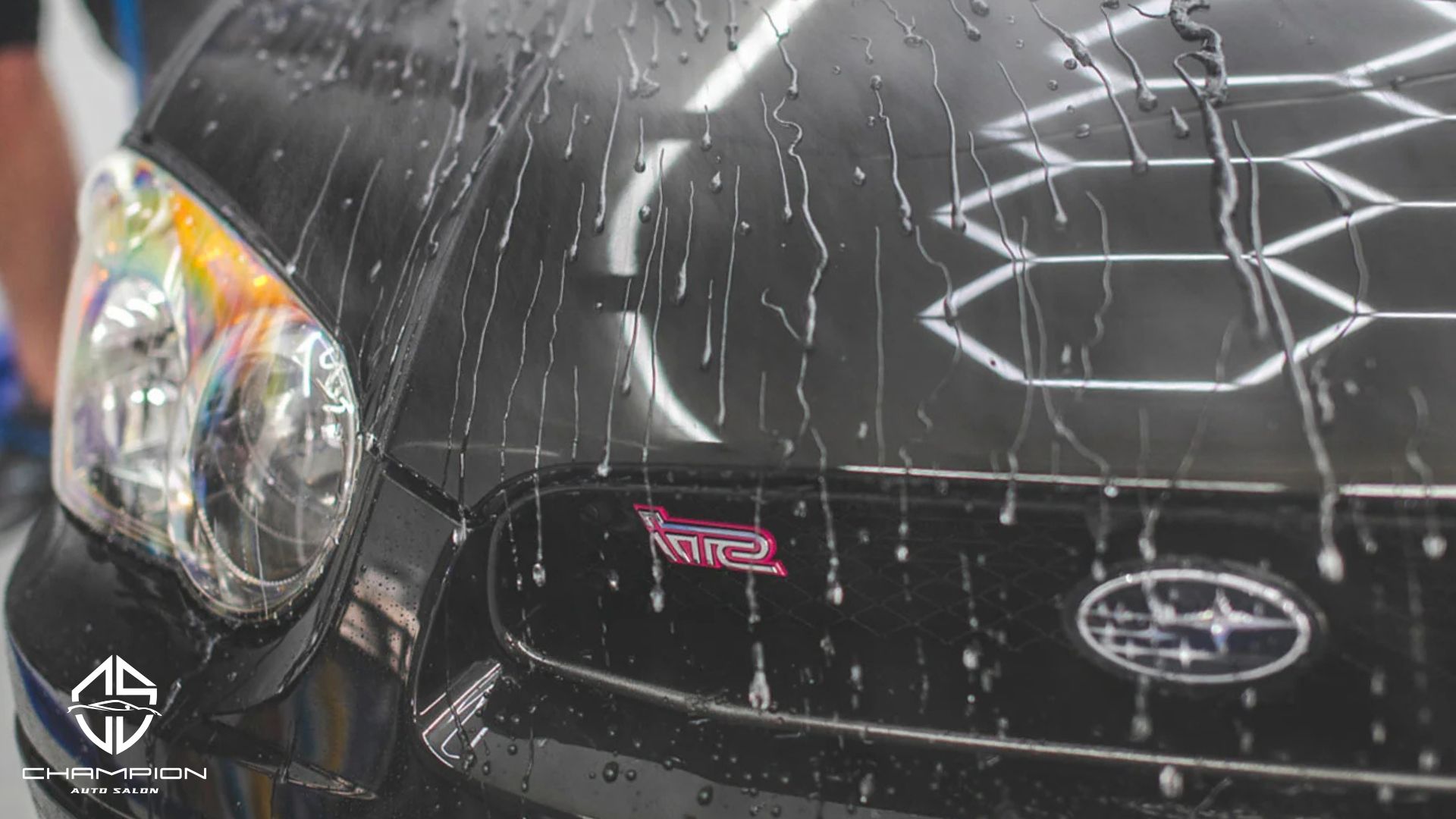 Water beads on the hood of a black Subaru WRX STI, showing the hydrophobic effect of ceramic coating. Champion Auto Salon logo is in the corner.