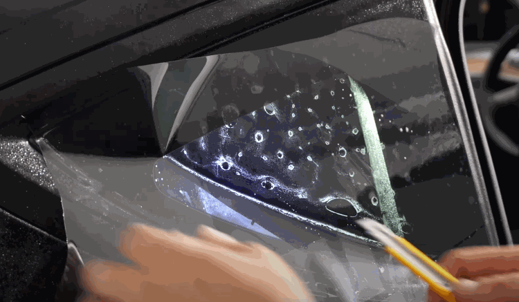 Close-up of hands applying windshield protection film to a car door window, with water and air bubbles visible under the film.