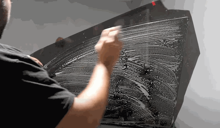 A person thoroughly cleans a large sheet of car glass using a sponge and soapy water in wide circular motions.